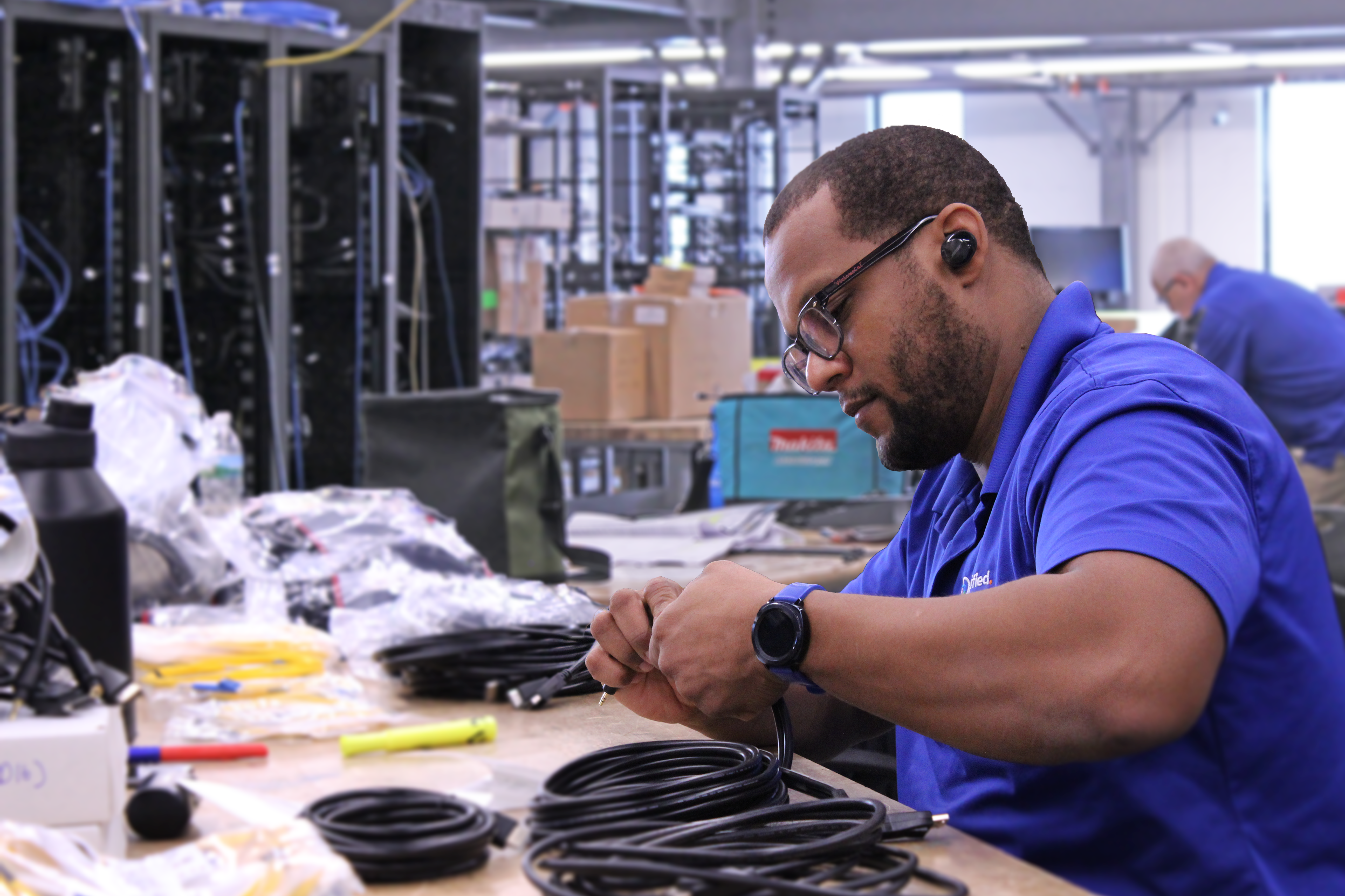 A man in a blue polo shirt works with cables at a table, wearing wireless earbuds and glasses.