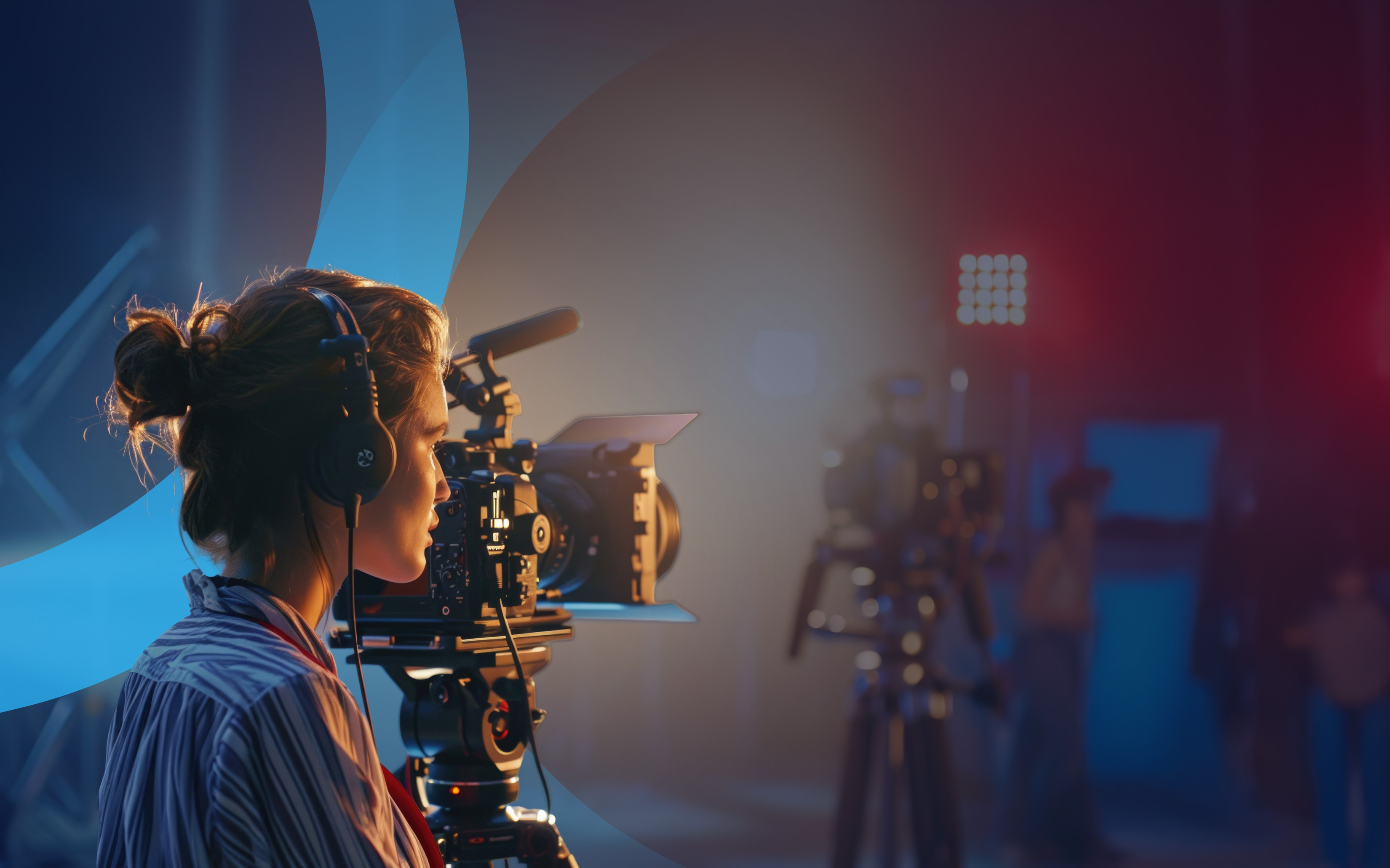 A woman operates a professional video camera on a film set with blue and red studio lights in the background.